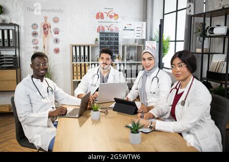 Gruppe von vier internationalen Ärzten in weißem Laborkittel, die zusammen am Tisch sitzen und moderne Laptops und Tablets verwenden. Medizinische Konferenz von männlichen und weiblichen Spezialisten. Mit Wissen teilen. Stockfoto