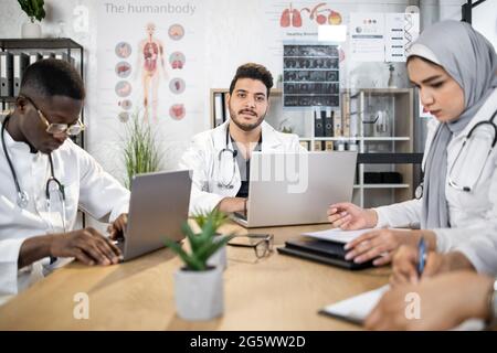 Vielfältiges medizinisches Team in weißen Laborkittel mit Notfallsitzung im Büro. Vier Spezialisten, die moderne Geräte im Raum einsetzen. Konzept der Zusammenarbeit und Diagnose. Ein arabischer Mann schaut in die Kamera Stockfoto