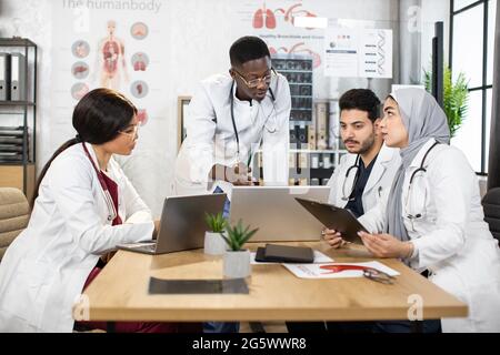 Vier verschiedene medizinische Mitarbeiter in Uniform, die im Büro mit modernen Geräten auf dem Tisch diskutieren. Konzept der Gesundheitsfürsorge und Teamarbeit. Stockfoto