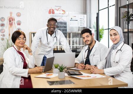 Multirassische Gruppe von Ärzten, die weiße Laborkittel und Stethoskope am Hals tragen, sitzen zusammen am Tisch und lächeln vor der Kamera. Medizinische Spezialisten während der Sitzung im Krankenhaus. Kooperationskonzept. Stockfoto