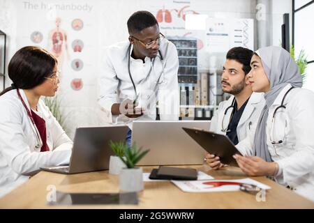 Vier verschiedene medizinische Mitarbeiter in Uniform, die im Büro mit modernen Geräten auf dem Tisch diskutieren. Konzept der Gesundheitsfürsorge und Teamarbeit. Stockfoto