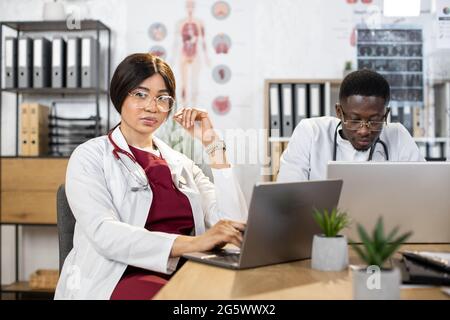 Multirassische Kollegen in medizinischen weißen Mänteln, die während der Konferenz im Krankenhaus an Laptops und Tablets arbeiten. Konzept der Gesundheitsversorgung, Technologie und Zusammenarbeit. Hübsche afrikanische Frau posiert vor der Kamera Stockfoto