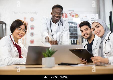 Internationales Team von kompetenten Ärzten in weißen Laborkittel mit modernen Geräten für dringende Treffen im Konferenzraum. Vier Kollegen analysieren gemeinsam unbekannte Viren. Stockfoto