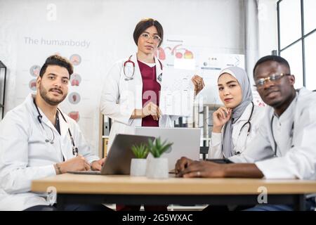 Professionelle Fachärzte, die während der Konferenz im Krankenhaus moderne Laptops und Tablets verwenden. Multirassische Ärzte diskutieren wichtige medizinische Themen im Sitzungssaal. Stockfoto