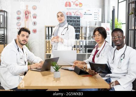 Vier verschiedene Menschen in medizinischen weißen Mänteln mit muslimischer Führerin, die mit modernen Geräten im Konferenzraum sitzt. Internationale Ärzte treffen sich im Krankenhaus. Konzept der Zusammenarbeit Stockfoto