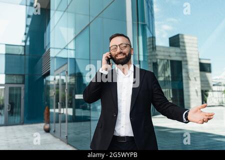 Geschäftsgespräch. Glücklicher, reifer Unternehmer, der auf dem Smartphone spricht und im Freien gegen das Bürozentrum steht Stockfoto