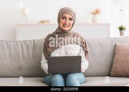 Glückliche Ältere Muslimische Dame, Die Zu Hause Einen Laptop Benutzt Und Hijab Trägt Stockfoto