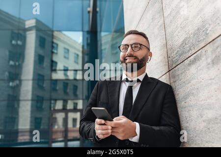 Glückliche Büroangestellte, die in der Nähe eines modernen Bürogebäudes mit einem Smartphone und Musik in kabellosen Kopfhörern hören Stockfoto