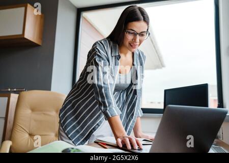 Charmante, fröhliche Frau, die mit einem Laptop arbeitet, während sie zu Hause am Tisch steht Stockfoto