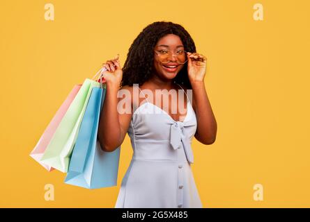 Saisonale Verkäufe und Shopping. Junge schwarze Frau trägt helle Einkaufstaschen mit Käufen, oranger Hintergrund Stockfoto