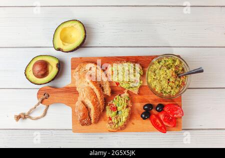 Flaches Foto, frisch zubereitete Guacamole in kleiner Glasschüssel, Brot, Tomaten, Oliven am Arbeitstisch und zwei Avocados am nächsten Schreibtisch aus weißem Holz Stockfoto