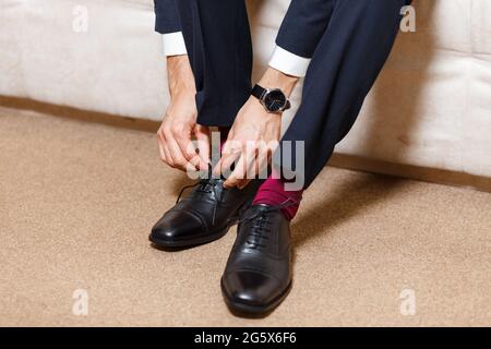 Ein Geschäftsmann in dunkelblauem Anzug, roten Socken und Uhren, der seine Schnürsenkel mit schwarzen Schuhen bindet. Moderner, stilvoller und teurer Look für junge Männer Stockfoto