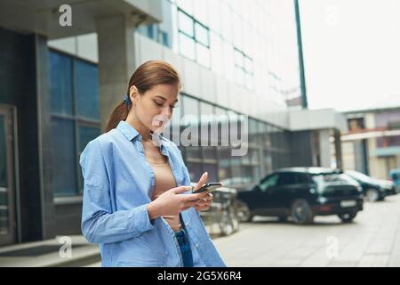 Weibliche Managerin nutzt in der Stadt im Freien Mobiltelefon, Frauen in blauem Hemd und Jeans schreiben eine Nachricht auf das Handy. Glückliche Geschäftsfrau, die am Telefon chattet. Stockfoto