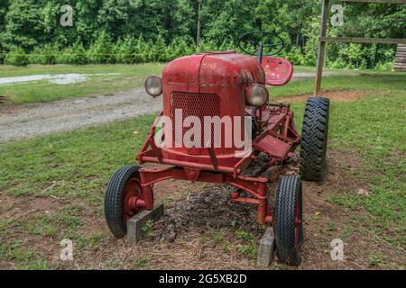 Alte alte alte kleine rote Bauernhof Traktor nicht mehr in Gebrauch geparkt auf Ackerland für die Anzeige im Freien mit Bäumen im Hintergrund Nahaufnahme Stockfoto