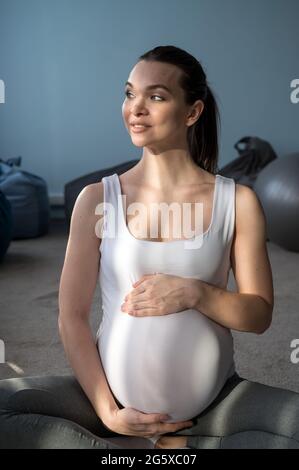 Ein trächtiges Mädchen sitzt in einer Lotusposition und hält sich an ihrem Bauch fest. Eine Frau macht Yoga im 3. Trimester. Eine blubbernde Mutter wartet. Stockfoto