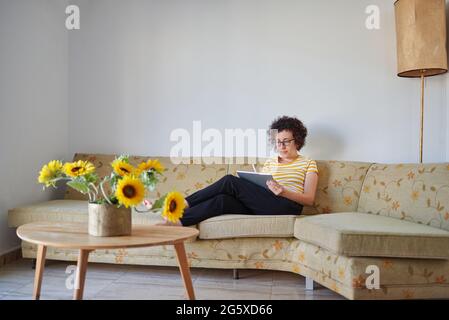 Die junge Frau greift auf ihr digitales Tablet, das auf dem Sofa sitzt Stockfoto