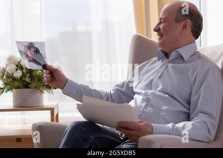 Allein ein älterer Mann sitzt zu Hause auf dem Sofa und hält Fotos, die an Erinnerungen denken Stockfoto