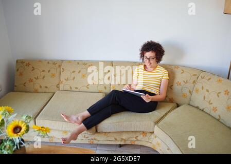 Die junge Frau greift auf ihr digitales Tablet, das auf dem Sofa sitzt Stockfoto