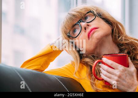 Porträt einer nachdenklichen oder besorgten erwachsenen Frau zu Hause, die mit einer Kaffeetasse aus dem Fenster blickt Stockfoto