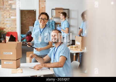 Mann und Frau lächeln vor der Kamera und arbeiten gerne gemeinsam an einem Spendenprojekt in Innenräumen. Sie berechnen, sortieren und verpacken Artikel in Stockfoto