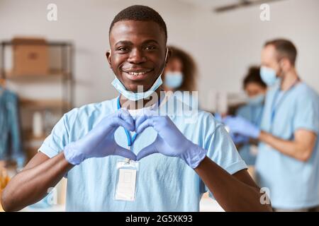 Nahaufnahme Porträt eines jungen afroamerikanischen Freiwilligen in blauer Uniform, Schutzmaske und Handschuhen, die die Kamera anlächeln und ein Zeichen der Herzliebe zeigen Stockfoto