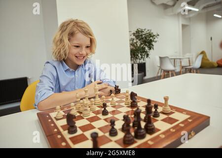Schachfan. Motivierter kaukasischer Junge, der aufgeregt beim Schachspielen am Tisch in der Schule sitzt Stockfoto