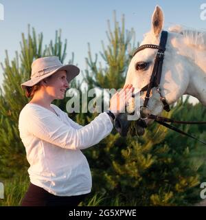 Eine junge Schwangerin füttert ein weißes Pferd auf dem Feld Stockfoto
