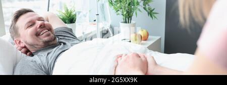 Lächelnder Mann liegt auf dem Krankenhausbett, seine Hand wird gehalten Von Frau Stockfoto