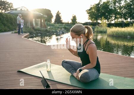 Online-Workout. Fröhliche, sportliche Frau winkt, während sie einen Videoanruf mit einem digitalen Tablet macht und an einem sonnigen Tag Yoga auf einer Matte in der Natur macht Stockfoto