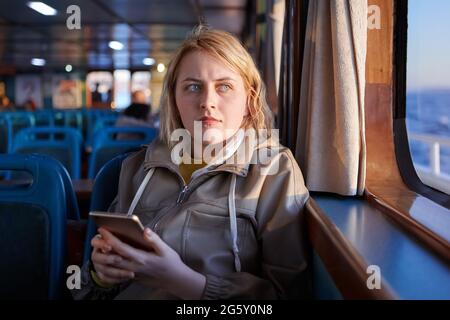 Traurige Passagierin der Fähre mit Handy in den Händen, schaut aus dem Fenster. Stockfoto