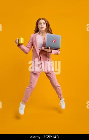 Besorgt nett Rotschopf erfolgreiche Frau Führer springen in der Luft tragen Laptop und Tasse Kaffee läuft schnell eilig Treffen isoliert über gelbe Farbe Stockfoto