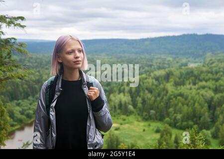 Das Mädchen im Teenageralter mit Rucksack geht auf dem Gipfel des Berges vor dem Hintergrund der bewaldeten Hügel und des Flusses spazieren Stockfoto