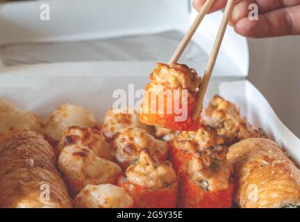 Hand hält eine Rolle mit Essstäbchen. Lunchboxen, gebackenes Sushi-Set. Verschiedene Brötchen auf hellem Hintergrund. Lieferung im Restaurant, Konzept zum Mitnehmen. Japane Stockfoto