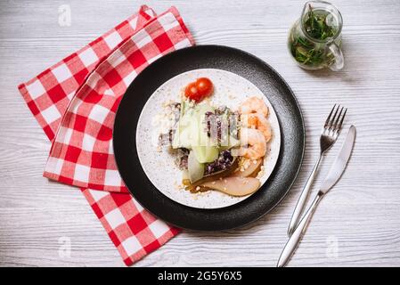Salat mit Flambe-Garnelen, Scheiben frischer Gurken und einer betrunkenen Birne, mit Kirschtomaten. Auf einem Holztisch mit Gabel und Messer, und einer Serviette. Stockfoto