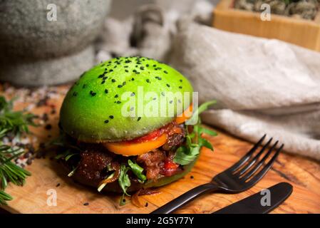 Hausgemachter Burger aus grünem Teig. Grünes Brötchen mit karamellisierten Zwiebeln, einer Scheibe Orangentomate, mit Rucola-Blättern, Barbecue-Sauce, gegrilltem Fleisch. Stockfoto