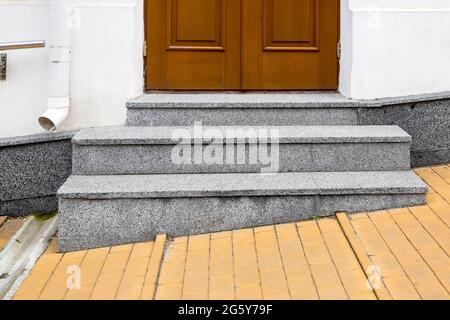 Granitschwelle an der Eingangstür aus braunem Holz und weißer Fassade mit Steinverkleidung des Gebäudes und Sturmrohrentwässerungssystem Vorderansicht, Stockfoto