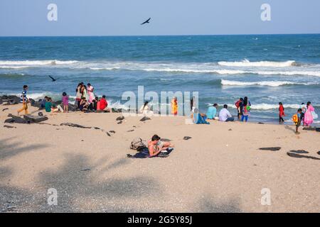 Westlicher männlicher Tourist mit rot sonnenverbrannter Haut sonnt sich am Strand mit Mobile, während indische Familien auf dem Seeweg spielen, Puducherry (Pondicherry), Tamil Nadu, Indien Stockfoto