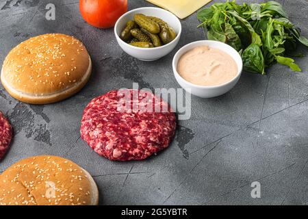 Zutaten zum Kochen von Burgern. Hacksteaks, Brötchen, Tomaten, Kräuter und Gewürze, auf grauem Steinhintergrund, mit Platz für Text Stockfoto