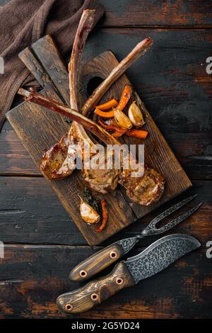 Hausgemachte gekochte Lammkoteletts mit Knoblauch- und Karotten-Set, auf einem hölzernen Servierbrett, auf einem alten dunklen Holztisch, von oben gesehen flach liegend Stockfoto