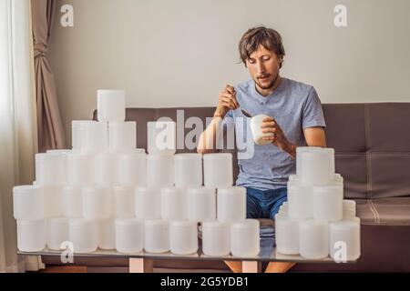 Der Mann isst Joghurt und ist von vielen Joghurtgläsern umgeben, die er in einem Jahr verwendet. Eine riesige Menge von Plastikbehältern für Lebensmittel. Übermäßiger Verbrauch von Stockfoto