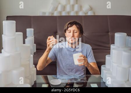 Der Mann isst Joghurt und ist von vielen Joghurtgläsern umgeben, die er in einem Jahr verwendet. Eine riesige Menge von Plastikbehältern für Lebensmittel. Übermäßiger Verbrauch von Stockfoto