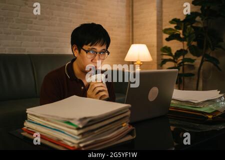 Gestresster asiatischer Geschäftsmann in legerer Kleidung, der bis spät in die Nacht mit Akte und Laptop arbeitet, während er Kaffee trinkt, um wach zu bleiben und sich zu Hause zu konzentrieren Stockfoto