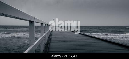 An einem düsteren Regentag in Schwarz und Weiß ragt ein einsamer, einsamer Pier in ein launisches Meer Stockfoto