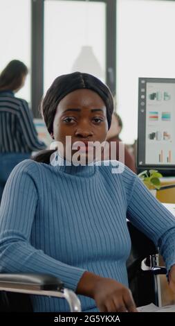 Junge schwarze Frau mit Behinderungen, ungültig, behindert Gehbehinderte, die die Kamera schauend verängstigt, traurig sitzend im Rollstuhl im Bürozimmer, beim Finanzprojekt mit dem Team arbeitend. Stockfoto