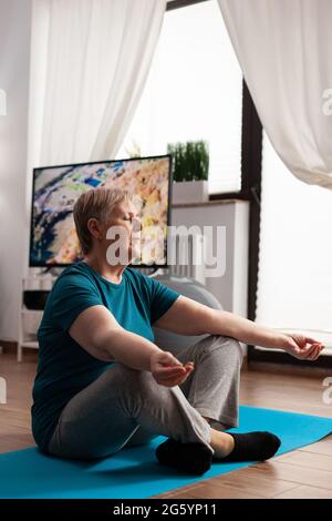 Friedliche ältere Frau, die bequem in Lotusposition auf einer Yogamatte sitzt und mit geschlossenen Augen Meditation praktiziert. Ruhiger Rentner, der Fitness trainiert, Körperübungen während des Meditationstrainings im Wohnzimmer Stockfoto
