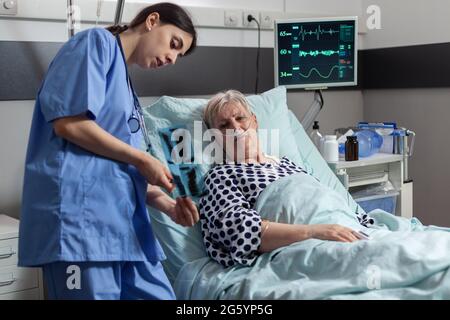 Alte Patientin mit Lungenerkrankung, die mit einer im Krankenhausbett liegenden Sauerstoffmaske atmet, lauschende Krankenschwester, die vor der Operation eine Röntgendiagnostik zeigt. Behandlung durch intravenöse Leitung. Stockfoto