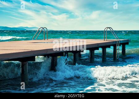 Hölzerner Pier mit Metallleitern, plätschernden Wellen und dem Meer an einem windigen Sommertag. Stockfoto