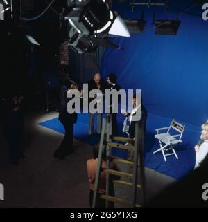 Telly Savalas, amerikanischer Schauspieler, Titeldarsteller der Krimi-Serie Kojak, zu Gast im ZDF, Musiksendung Disco, 1975, hier bei einer Studioprobe mit Moderatorin Ilja Richter und Fotograf. Telly Savalas, amerikanischer Schauspieler, Titelfigur der Krimiserie Kojak, Gast im ZDF, Musikshow-Disco, 1975, hier bei einer Studioprobe mit Moderatorin Ilja Richter und Fotografin. Stockfoto