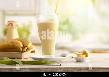 Milchshake mit Banane in Glas Tasse auf Tisch mit Tablett mit Obst und Küche Hintergrund. Vorderansicht. Stockfoto