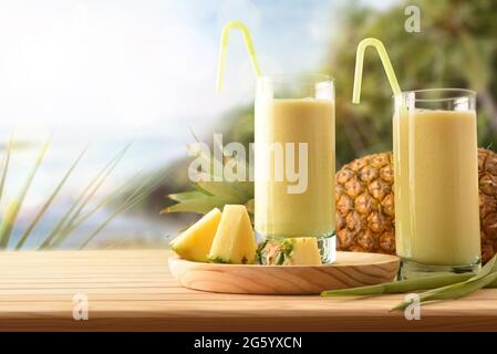 Zwei große Gläser mit Ananassaft und Milch- und Ananasportionen auf einem Holztisch mit tropischem Küstenhintergrund. Vorderansicht. Stockfoto